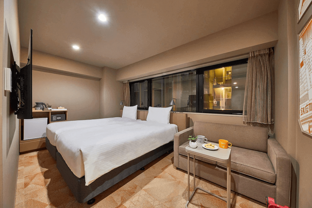 lyf銀座東京舒適雙床房，房內配置兩張單人床與一張實用的淺褐色雙人沙發及小茶几，大面窗戶引入明亮採光，整體空間呈現溫馨且現代的實用設計風格。