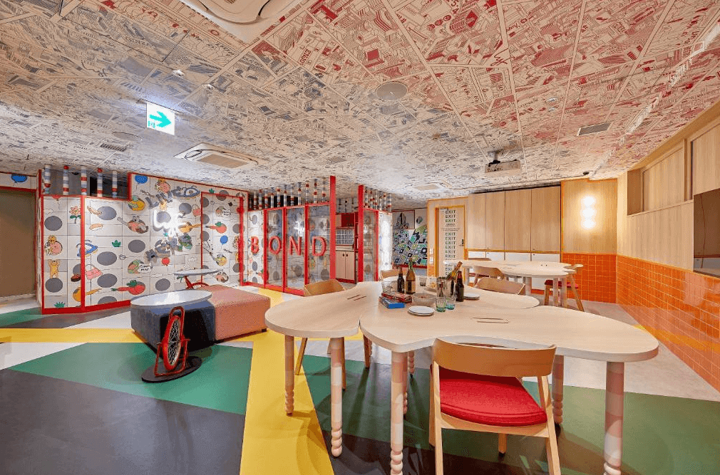 lyf銀座東京名為「BOND」的公共交誼廳，擁有色彩繽紛的幾何拼色地板與不規則造型的木質大桌，天花板延續手繪地圖元素，為旅人提供了一個充滿活力與靈感的社交及辦公空間。