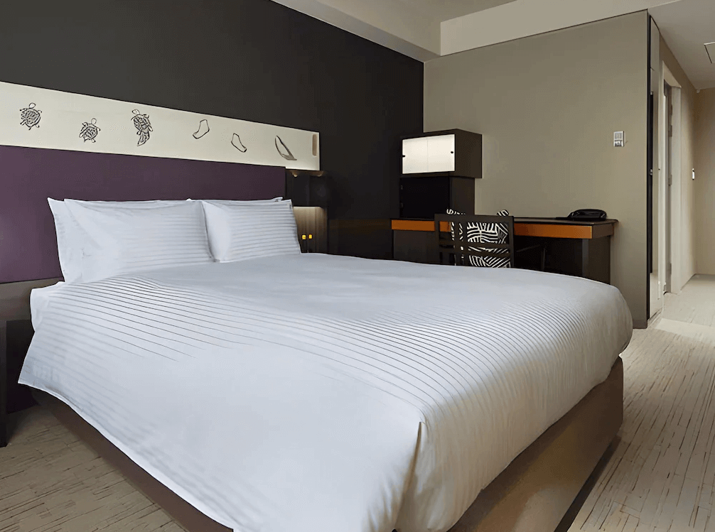 三井花園飯店京橋東京站的舒適雙人房，床頭背板採用獨特的深紫色搭配充滿趣味的日式傳統圖騰插畫，配備純白大床與質感深色木紋辦公桌，展現日式摩登與現代簡約交織的都會風格。