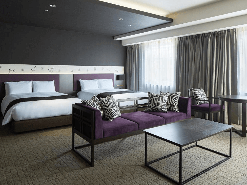 三井花園飯店京橋東京站的寬敞雙床客房，房內配置兩張舒適大床，並在床尾處設有質感的深紫色長型沙發與深色茶几，大面窗戶引進明亮採光，整體空間設計沉穩且大氣。