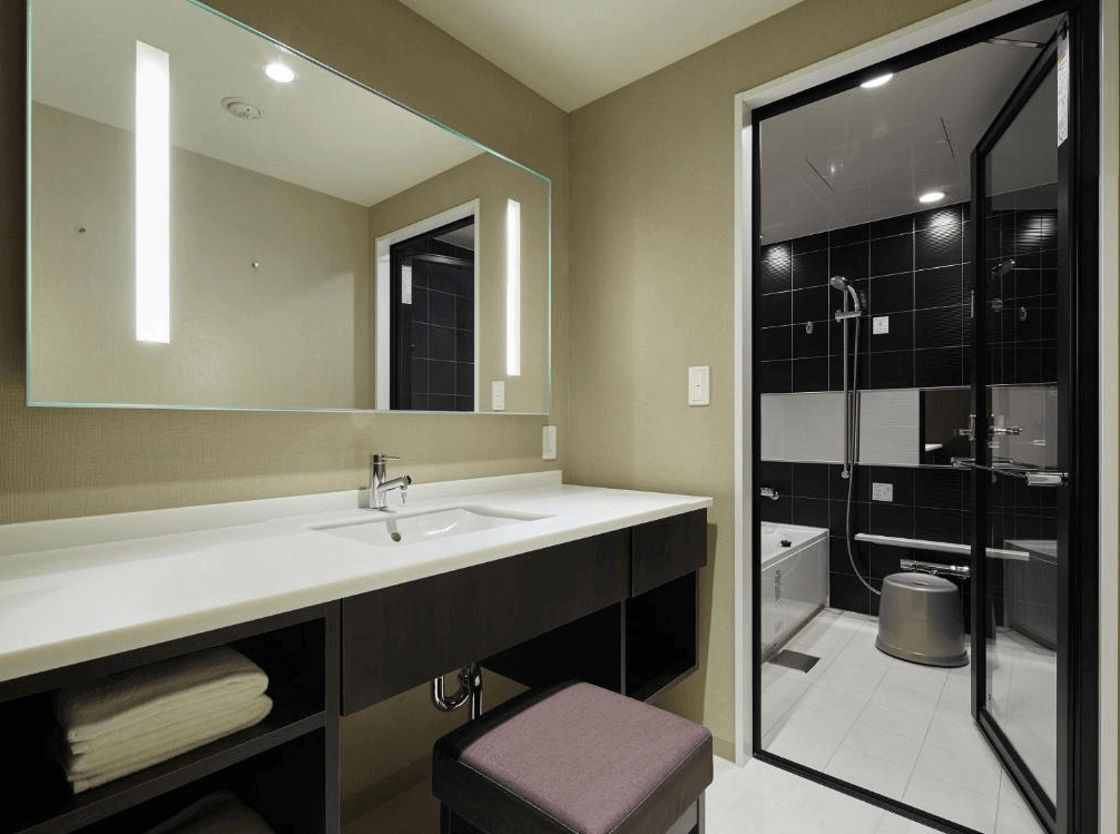 三井花園飯店京橋東京站的乾濕分離衛浴空間，外側設有寬敞明亮的白色洗手檯面與大面梳妝鏡；內側則為採玻璃門設計的日式獨立洗浴空間，牆面鋪設深黑色磁磚，極具現代洗鍊質感。