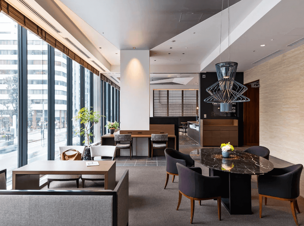 三井花園飯店京橋東京站的質感公共大廳與休息區，擁有大面落地窗可直接欣賞繁華的都會街景，室內配置深色大理石橢圓桌、充滿前衛設計感的幾何造型吊燈與舒適的沙發座位，營造出高級且寧靜的空間氛圍。