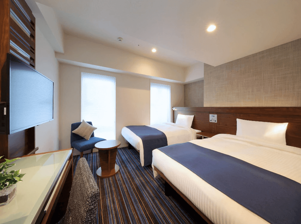 京王布萊索飯店東京站八重洲舒適雙床房，房內採用沉穩的深藍色床尾巾與同色系單人沙發椅，搭配溫潤的木質床頭板與條紋地毯，空間簡約舒適且自然採光良好。