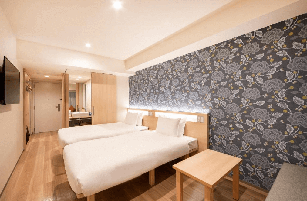 唐草飯店東京站的標準雙床客房，房間採用明亮的淺色木質地板與溫潤家具，床頭主牆貼有優雅的深灰色花卉圖騰壁紙，為簡約的現代空間增添了一抹沉穩的設計質感。