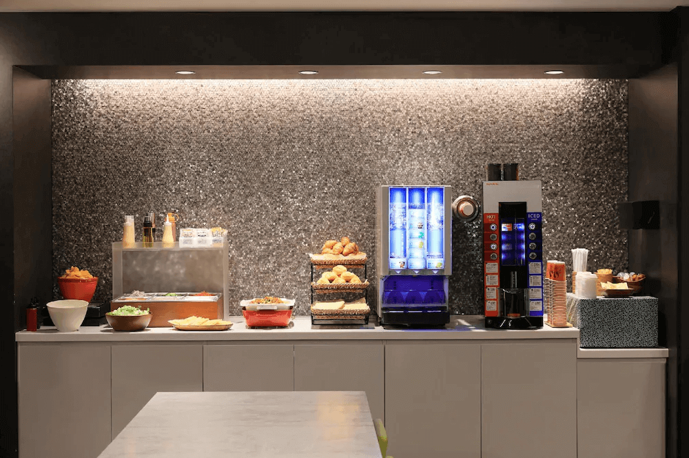 唐草飯店東京站的公共自助休息區，吧台背景採用閃亮的灰色馬賽克磁磚裝飾，檯面上整齊擺放著各類麵包輕食，並設有多功能的冷熱飲水機與咖啡機，提供旅客便利的餐飲服務。