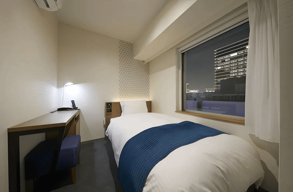 東京京橋穎特飯店的簡約單人房，配有舒適的單人床與深藍色床尾巾，靠牆設有結合收納功能的實用木質書桌，大面窗戶可飽覽繁華的東京都會夜景，空間精巧且具現代感。