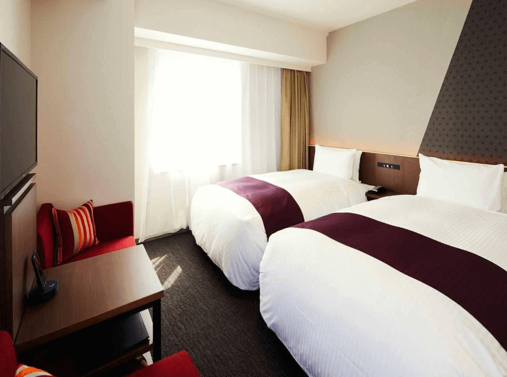 東京京橋穎特飯店的舒適雙床房，床鋪點綴著優雅的酒紅色床尾巾，角落配置了一張搶眼的紅色單人沙發與木質小茶几，大面窗戶引入明亮的自然採光，住宿氛圍溫馨放鬆。