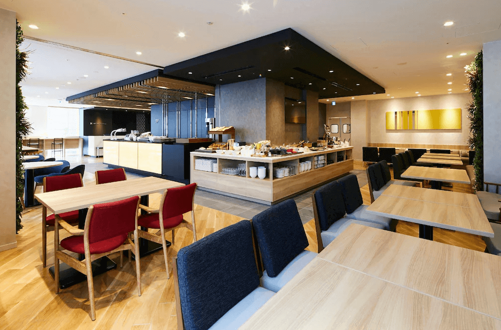 東京京橋穎特飯店的公共餐廳與自助吧台區，採用溫潤木質地板搭配跳色的紅、藍休閒座椅，中央的中島取餐動線寬敞流暢，提供旅客舒適明亮且充滿活力的用餐環境。