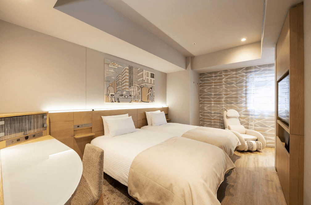 東京京橋雷姆飯店精緻雙床房，床頭牆面掛有描繪都市街景的藝術畫作，增添都會氣息，房內同樣貼心配置了紓壓按摩椅與質感休憩小桌，整體設計簡約明亮且具現代感。