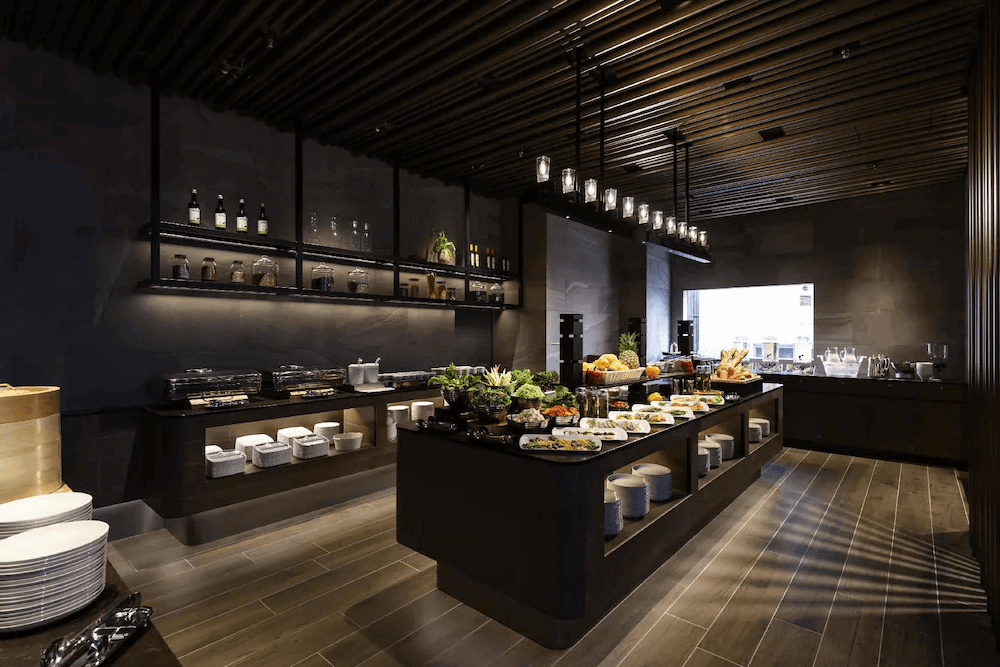東京京橋雷姆飯店的自助式早餐吧台，採用沉穩的深灰色石材與深色木質裝潢，搭配時尚的玻璃垂吊燈飾，中島檯面上豐盛地陳列著各式生菜沙拉與冷熱食，用餐環境充滿都會洗鍊質感。
