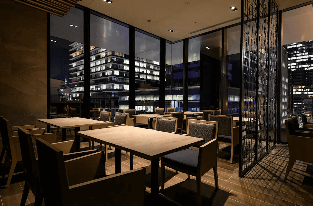 東京京橋雷姆飯店高空景觀餐廳座位區，室內配置深色木質餐桌與舒適的布面單椅，透過環繞式的大面落地窗，可將繁華璀璨的東京大樓夜景盡收眼底，氛圍極致浪漫迷人。