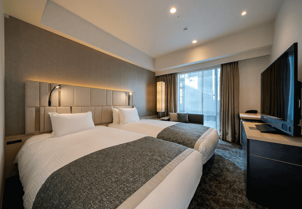 東京京橋大及魯內普米爾飯店舒適雙床房，床頭採用溫潤的高質感軟墊與專屬閱讀燈，房內配置大型液晶電視與靠窗的沙發休憩區，整體空間設計沉穩且典雅。