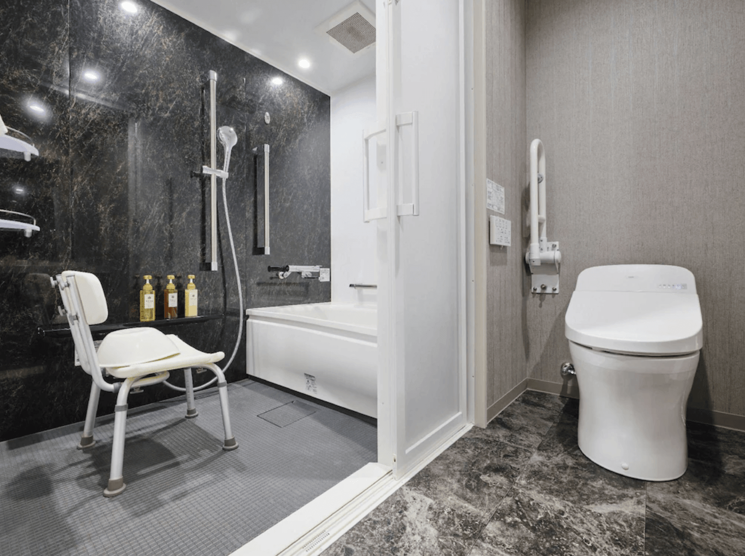 飯店內部的無障礙衛浴設施。浴室採用灰黑色大理石紋理牆面，設有浴缸、淋浴區、專用助浴椅以及備有安全扶手的免治馬桶空間
