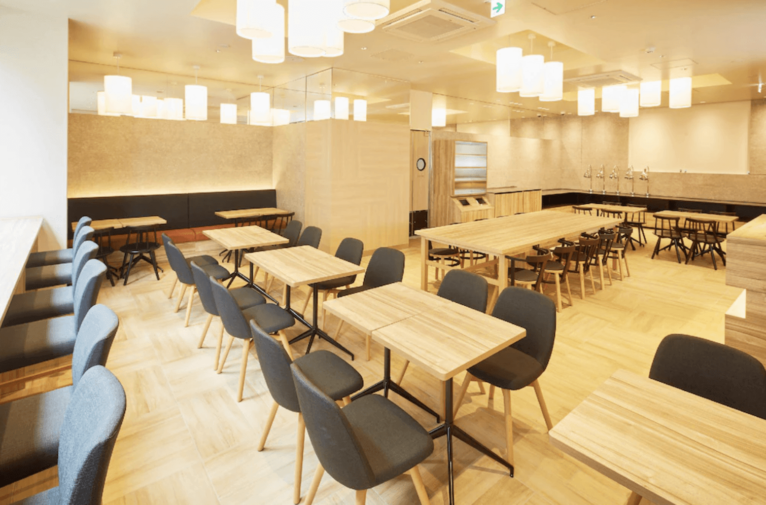 設計簡約明亮的飯店餐廳用餐區。室內擺放多組木質長桌與簡約座椅，搭配天花板上錯落有致的圓柱形白色吊燈，營造出溫馨舒適的用餐氛圍