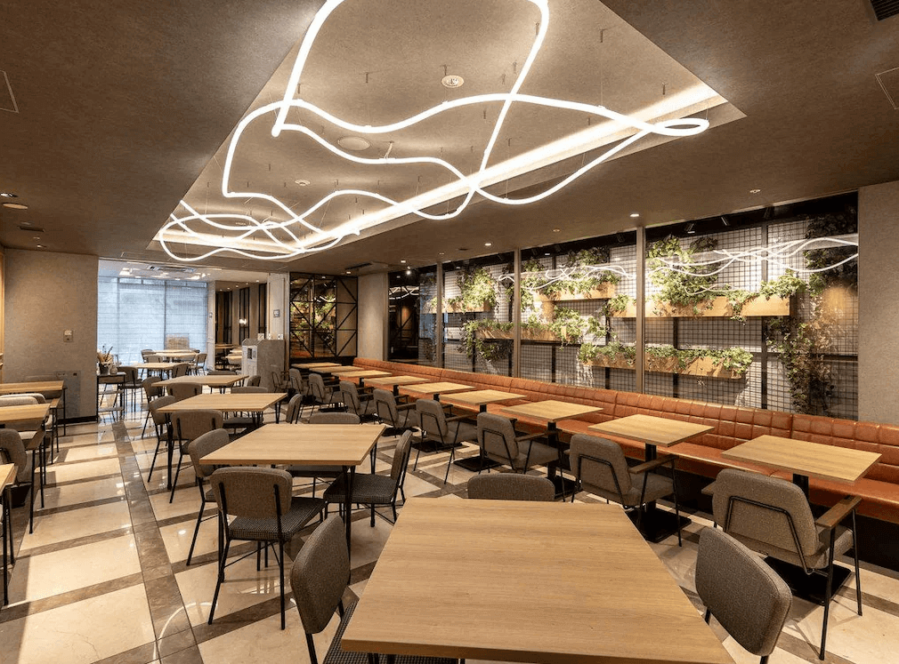 東京站八重洲中央口超級精選飯店寬敞的公共餐廳與休憩交誼區，天花板設有極具現代設計感的流線型造型燈飾，背牆以鐵件網格結合綠色植栽打造出城市綠洲般的視覺焦點，配置充足的木質桌椅與舒適的橘色長沙發。