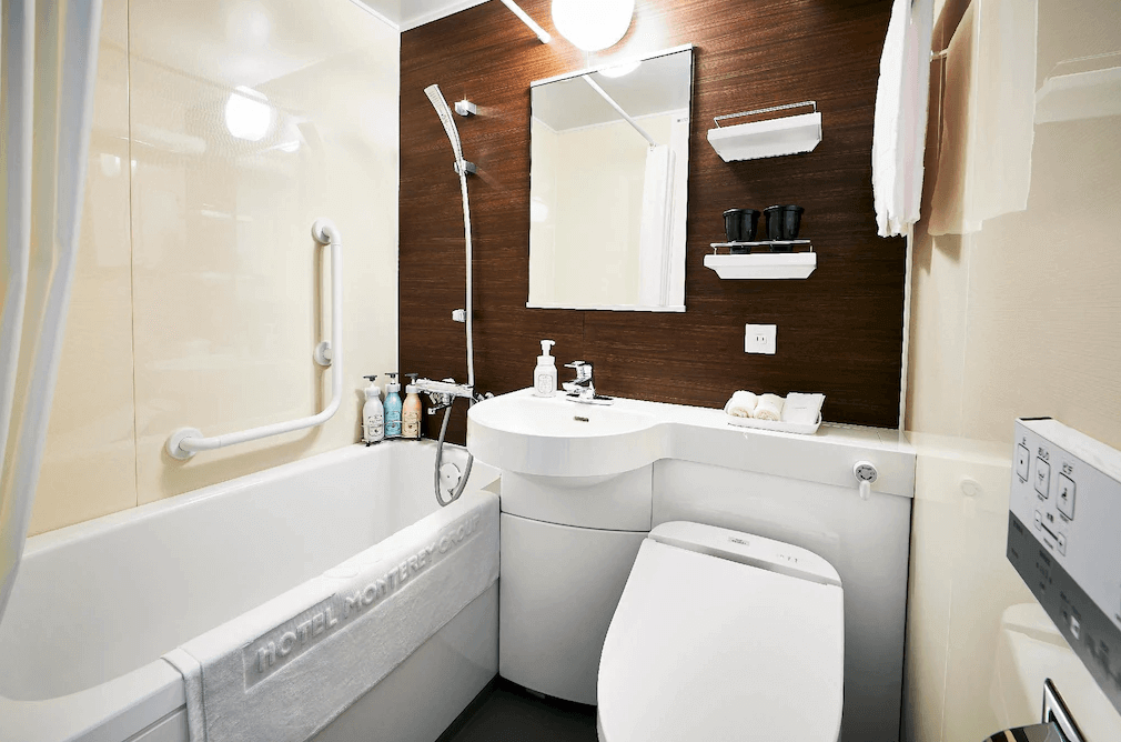 東京蒙特埃爾馬納飯店明亮整潔的衛浴空間，洗手台背牆特別採用深色木紋飾板增添設計層次，配備免治馬桶、洗沐備品與附設安全扶手的舒適浴缸，提供旅客放鬆的盥洗環境。