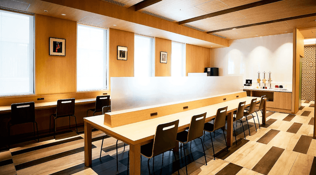 東京蒙特埃爾馬納飯店公共休息區，空間以大量的淺色木質裝潢為主，中央設有配置霧面隔板的長型共用辦公桌，後方備有自助茶水吧台，環境寬敞明亮，適合旅客在此辦公或小憩。