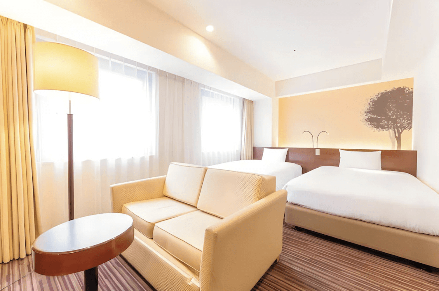 淺草京阪飯店 (Hotel Keihan Asakusa) 採光明亮的客房空間，設有兩張舒適單人床、米色沙發與大型落地燈，牆面裝飾著簡約的樹木圖騰，環境典雅舒適