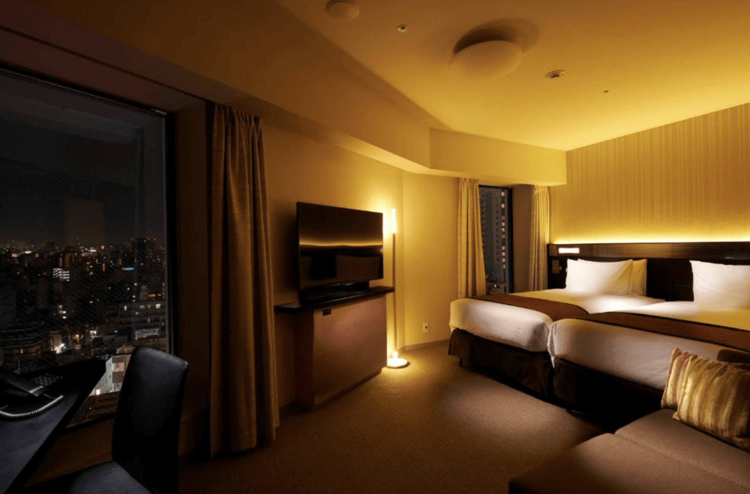 里士滿淺草國際酒店三人房（沙發床房）的夜間實景。房間燈光溫馨，配有兩張單人床和一張沙發床，以及長書桌、電視和落地燈。大片落地窗外展現迷人的淺草城市夜景