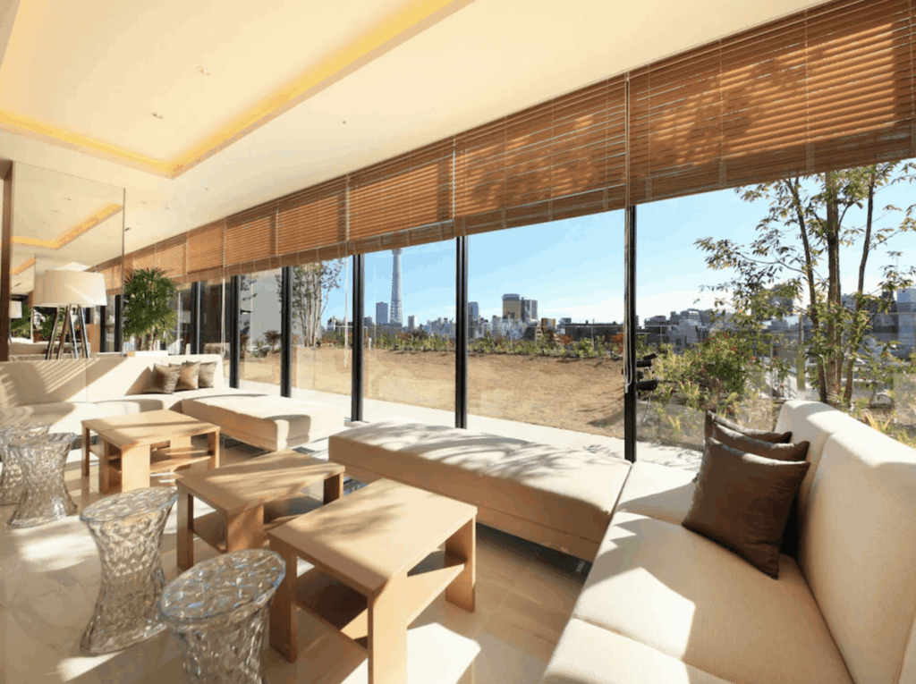 "淺草住宿精選示意圖"里士滿淺草國際酒店的公共休息室。大片落地窗帶來充足的自然光，配有舒適的米色沙發、木質和透明小邊桌。窗外是戶外草坪區，遠處清晰可見東京晴空塔矗立在蔚藍天空下