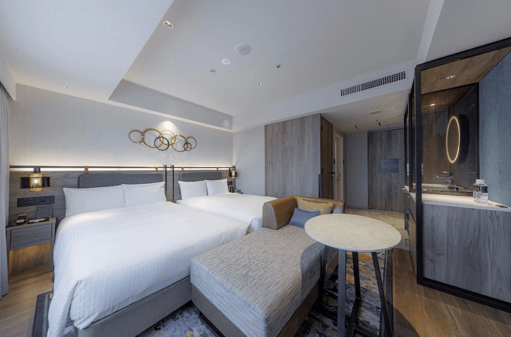 JR九州小倉車站飯店 (JR Kyushu Station Hotel Kokura)1.jpg
飯店雙床房實景。房間採用現代簡約設計，配有兩張單人床與木質床頭板，上方裝飾有圓環藝術品。房內設有一張長型沙發床與大理石面圓几。洗手台位於睡眠區旁的開放式空間，整體動線開闊。