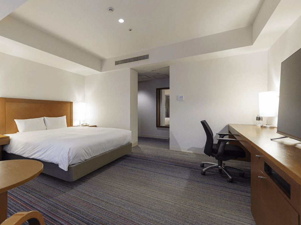 JR九州小倉車站飯店 (JR Kyushu Station Hotel Kokura)2.jpg 飯店單床房（大床房）實景。室內空間寬敞，鋪設條紋地毯。右側為大型木質辦公桌與黑色辦公椅，上方放置電視。房間色調溫馨，配有柔和的床頭燈與大面積留白牆面，營造出寧靜的商務休息空間。