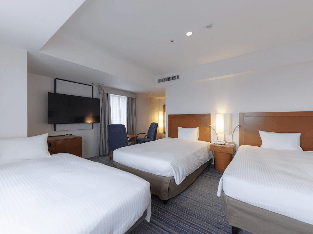 JR九州小倉車站飯店 (JR Kyushu Station Hotel Kokura)3.png 飯店三床房（三人房）實景。房內整齊擺放三張單人床，每張床皆有獨立的床頭靠板。窗邊設有藍色扶手椅與小桌。牆面上掛有大型液晶電視，非常適合家庭或團體旅客共同入住。
