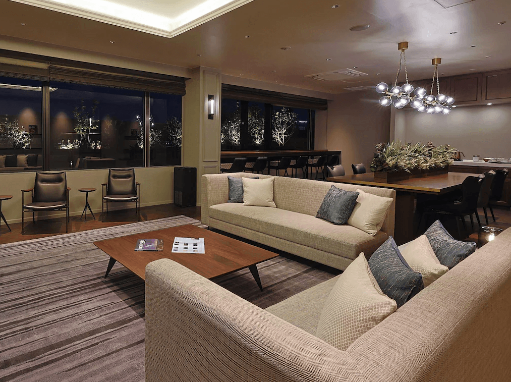 JR九州小倉車站飯店 (JR Kyushu Station Hotel Kokura)4.jpg 飯店的大廳休息區或行政廊。空間佈置典雅，設有大型淺色布藝沙發與胡桃木色咖啡桌。後方有長型餐桌、設計師燈飾以及景觀窗台位，窗外可見夜晚的綠化造景，氛圍高級且舒適。