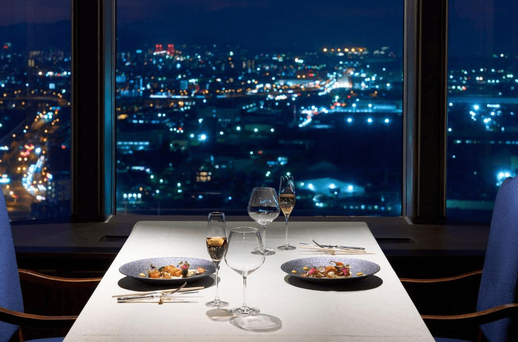 北九洲小倉麗嘉皇家飯店 (Rihga Royal Hotel Kokura Fukuoka)3.png 飯店餐廳的夜景晚餐場景。桌上擺放精緻的法式料理、香檳杯與水杯，背景是整片落地窗，呈現壯麗的北九州市中心夜景，燈火輝煌，氛圍浪漫且高級。