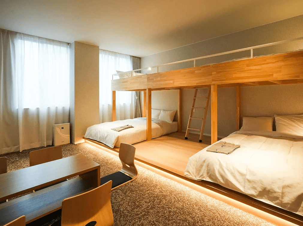 千草酒店 (Chigusa Hotel)2.jpg 飯店家庭房實景。房內設有獨特的兩層木製大通鋪，下層擺放兩張單人床墊，上層則有護欄。房間鋪設地毯，床位區下方有溫暖的間接照明。左側設有日式風格的矮桌與靠背椅，營造出適合多人入住的放鬆空間。