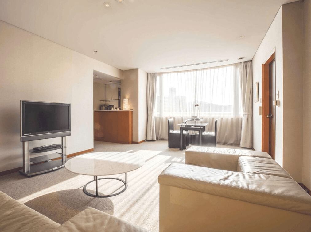 千草酒店 (Chigusa Hotel)4.jpg 飯店套房的客廳區域。空間非常寬敞，配有大型 L 型淺色皮質沙發、大理石面圓型茶几與液晶電視。落地窗前設有一組小型餐桌椅，陽光透過紗簾灑進室內，背景還可見簡易的吧檯區，整體設計大氣且採光極佳。