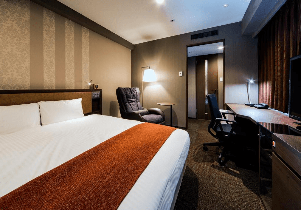 大和Roynet飯店 - 小倉站前 (Daiwa Roynet Hotel Kokura-Ekimae)2.jpg 飯店單人或雙人房實景。室內有一張寬敞的大床，床尾鋪著橘紅色床旗。房內配備一張深色按摩椅，右側長型辦公桌上擺放電視與電話。牆面採用優雅的條紋與花紋壁紙裝飾，營造溫暖舒適的氛圍。