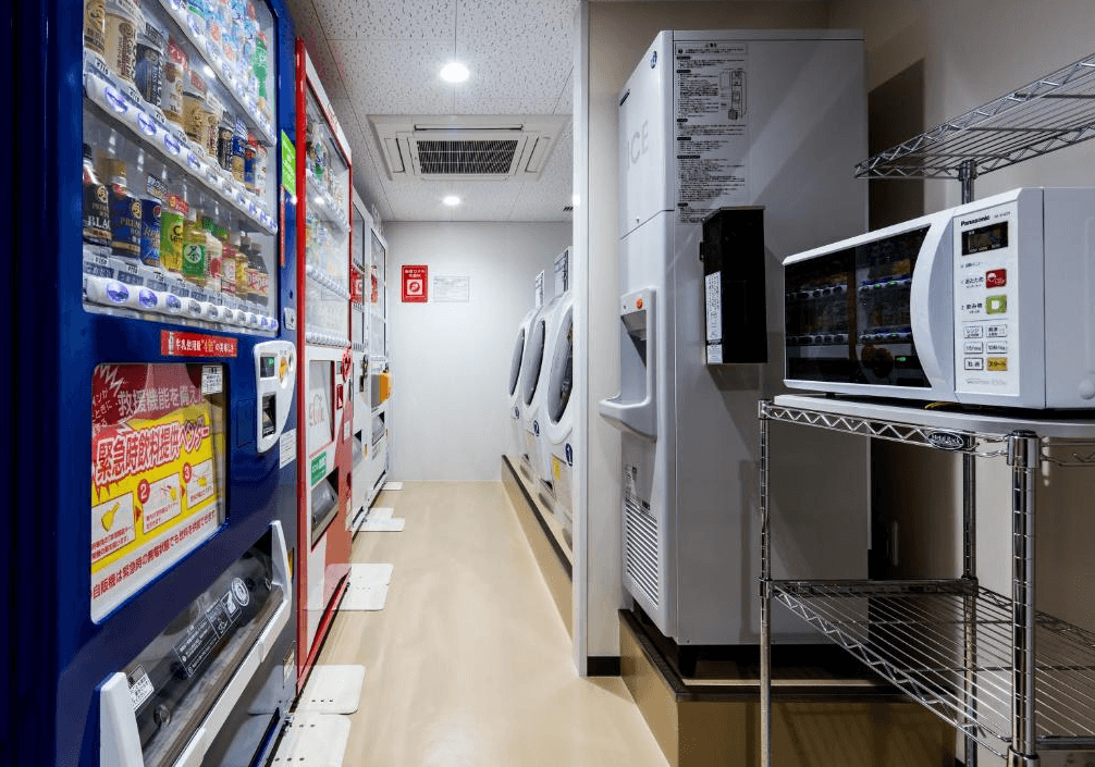 大和Roynet飯店 - 小倉站前 (Daiwa Roynet Hotel Kokura-Ekimae)4.jpg 飯店的公共自助設施區。窄長型的走廊內擺放著自動販賣機、洗脫烘一體滾筒洗衣機、製冰機，以及放置在層架上的微波爐，方便長住或有需求的房客使用。