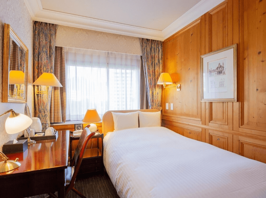 小倉新田川藝術酒店 (ART HOTEL Kokura New Tagawa)2.jpg 飯店單床房實景。房間採用經典的歐式木質裝潢，牆面鋪設精緻壁紙並掛有裝飾畫。房內配置一張單人床與深木色辦公桌，桌上設有典雅的檯燈與梳妝鏡。窗戶搭配花卉圖案的厚質窗簾，營造出溫暖且具歷史感的氛圍。