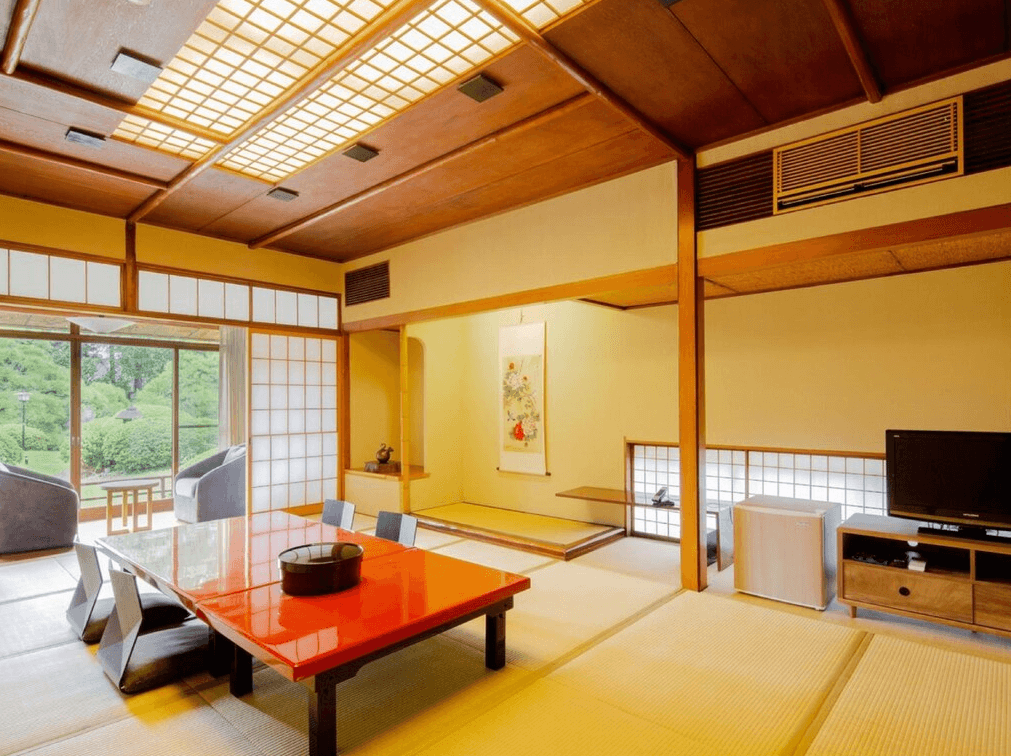 小倉新田川藝術酒店 (ART HOTEL Kokura New Tagawa)3.png 飯店傳統日式客房（和室）。室內鋪設榻榻米，設有紅色漆面矮桌與和式靠背椅。牆面掛有捲軸畫，天花板採用木質格柵與紙燈裝飾。拉門後方設有緣側休息區，可一邊喝茶一邊欣賞窗外的日式庭園景色。