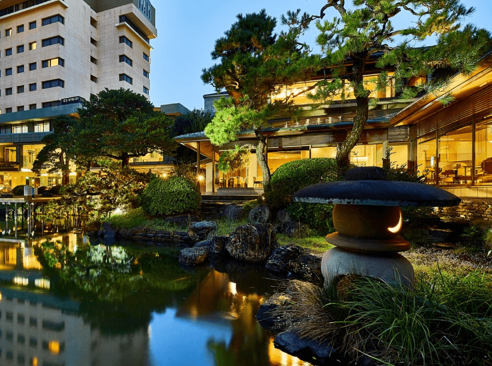 【北九州住宿精選示意圖】小倉新田川藝術酒店 (ART HOTEL Kokura New Tagawa)4.jpg 飯店著名的百年日式庭園夜景。前景有一座刻有月亮圖案的石燈籠，池塘倒映著建築的燈火。庭園內點綴著古松與翠綠植栽，後方則是現代化的飯店建築，展現出傳統與現代融合的獨特景致。