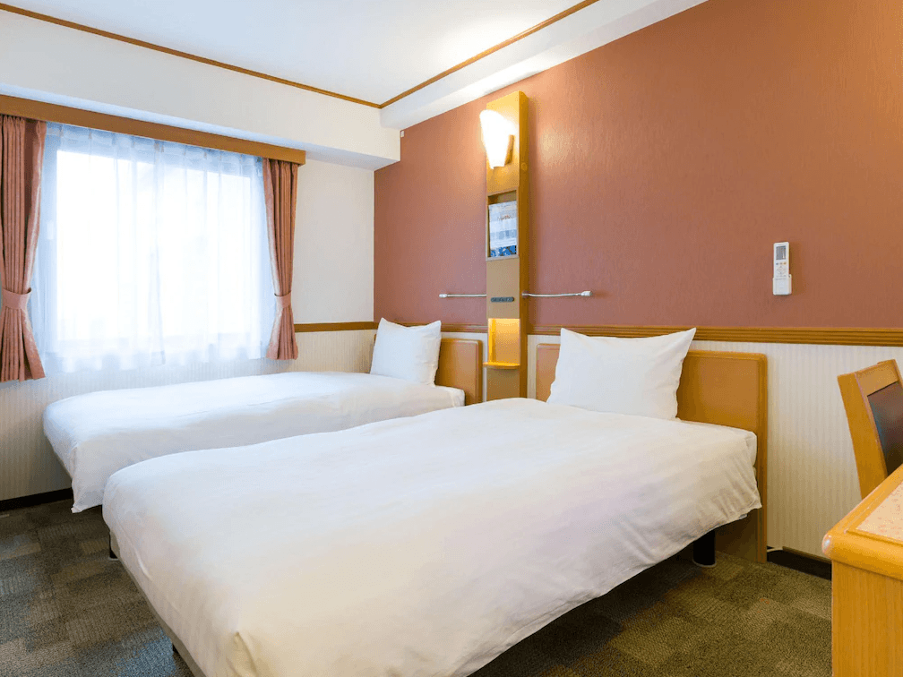 東橫INN北九州機場 (Toyoko Inn Kitakyushu Airport)1.png
飯店雙床房實景。房間採用東橫INN經典的溫馨風格，牆面為淡橘色調。房內配置兩張單人床，床頭設有整合式閱讀燈與收納架。窗戶配有粉色遮光窗簾，整體空間潔淨且採光良好。
