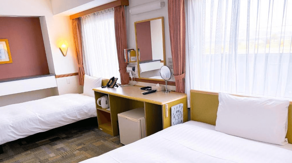東橫INN北九州機場 (Toyoko Inn Kitakyushu Airport)2.png 飯店雙床房的另一視角。兩張單人床之間擺放著長型木質書桌，桌上配備電話、鏡子與梳妝鏡，桌下則收納有電熱水壺與小冰箱。大片窗戶搭配白色紗簾，光線充足且舒適。