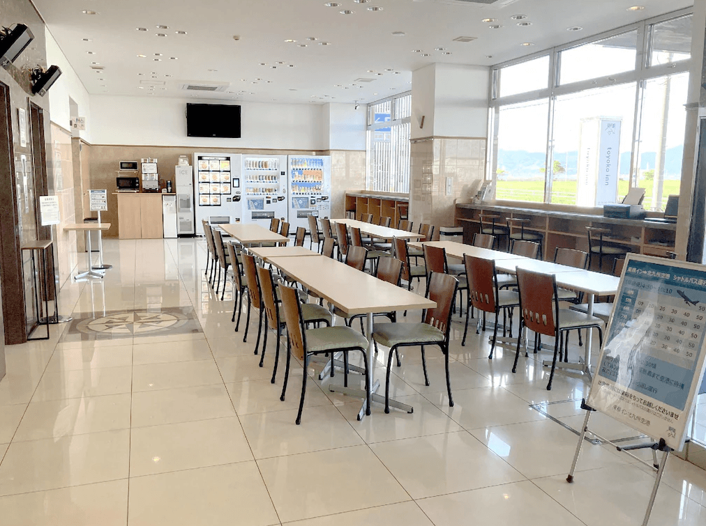 東橫INN北九州機場 (Toyoko Inn Kitakyushu Airport)3.jpg 飯店一樓的公共用餐與休息區。寬敞明亮的空間擺放多組木質長桌椅，右側是大面積落地窗，可以看見室外草地。背景設有自動販賣機、製冰機與微波爐區，入口處立有機場接駁巴士時刻表。