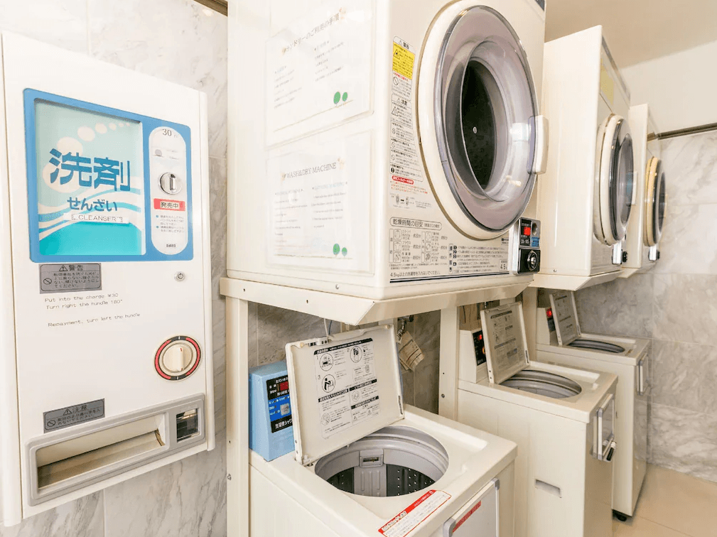 東橫INN北九州機場 (Toyoko Inn Kitakyushu Airport)4.jpg 飯店內的自助洗衣房設施。牆邊整齊排列著投幣式洗衣機與上方堆疊的烘衣機。左側牆面裝有洗衣粉自動販賣機。空間採用淺色大理石紋牆面，環境明亮且維護良好，適合長住旅客使用。