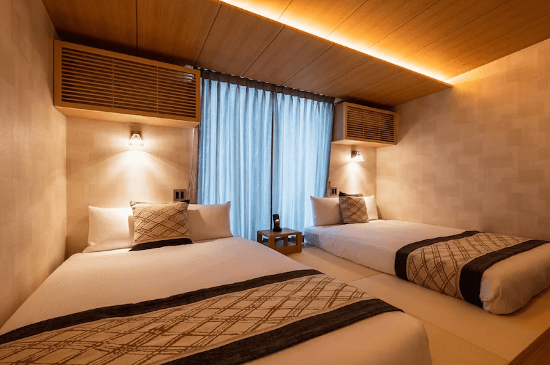 格蘭德貝塞門司港站前酒店 (GRAND BASE Mojiko Ekimae)1.png
飯店雙床房實景。房間採用現代和風設計，配置兩張並列的單人床。床頭上方有木質格柵裝飾與溫馨的壁燈，天花板設有間接照明。窗戶配有透光的藍灰色紗簾，整體營造出沉穩且放鬆的睡眠氛圍。