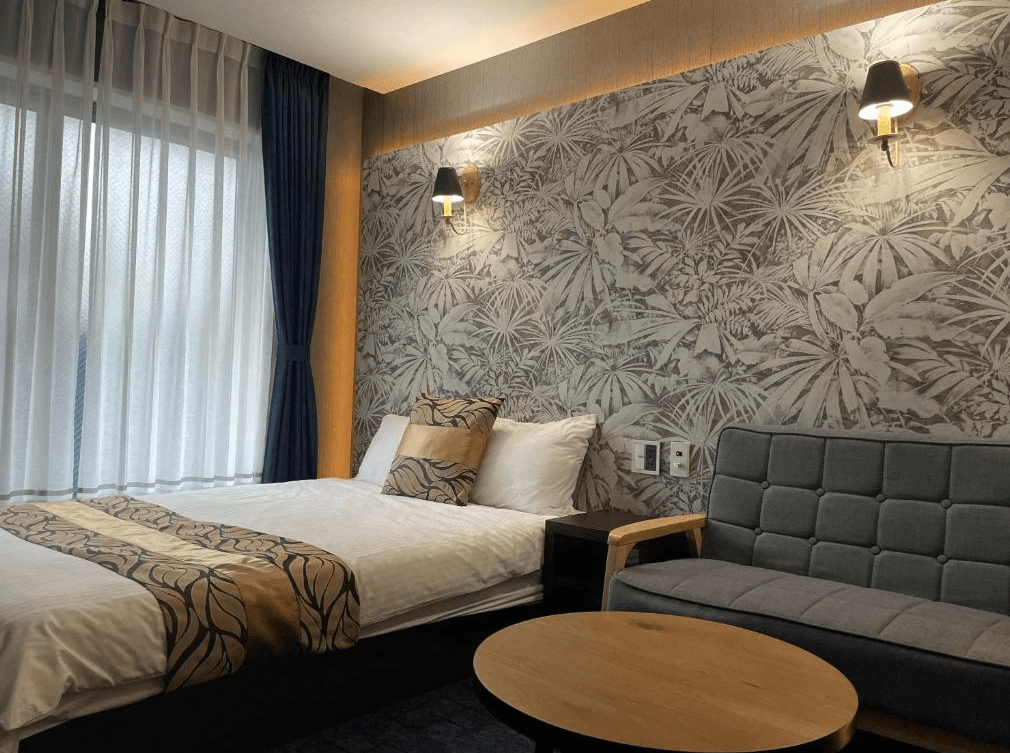 格蘭德貝塞門司港站前酒店 (GRAND BASE Mojiko Ekimae)2.jpg 飯店單人或雙人房實景。床頭牆面使用大面積的灰色熱帶植物圖騰壁紙，並配有兩盞黑色復古壁燈。床側設有一座灰色拉鈕造型沙發與木質圓形茶几，落地窗配有深藍色遮光簾與白紗簾，空間極具設計感。