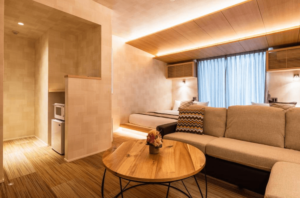 格蘭德貝塞門司港站前酒店 (GRAND BASE Mojiko Ekimae)3.png 飯店寬敞的起居空間。前景為大型淺色布質 L 型沙發與原木色茶几，背景可見架高的床位區。房間左側牆內嵌入了小型微波爐與冰箱，動線設計流暢，適合家庭或長期住宿旅客使用。