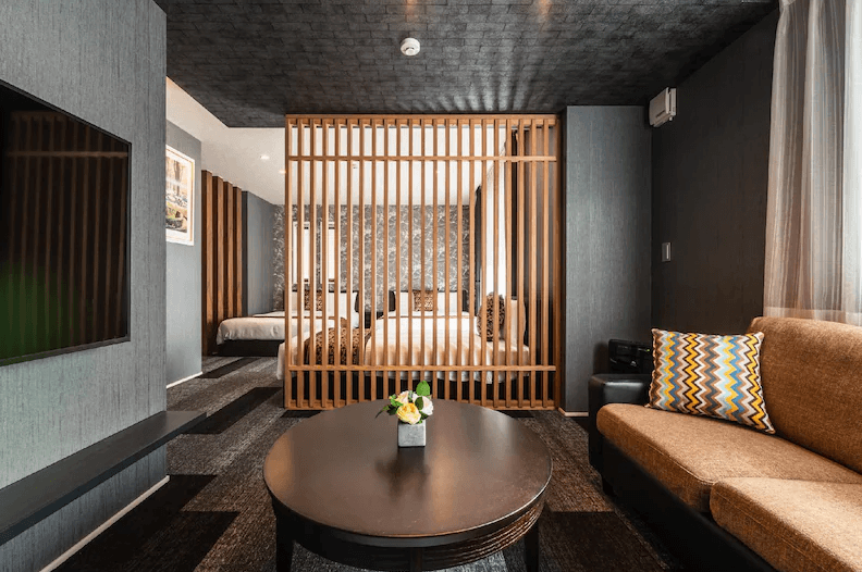 格蘭德貝塞門司港站前酒店 (GRAND BASE Mojiko Ekimae)4.png 飯店行政或豪華房型實景。房間中央以大型木質格柵屏風區隔睡眠區與起居區。起居區配有棕色沙發、圓形黑木茶几與大型掛牆電視。室內風格以深灰色調搭配木質元素，呈現出高級且具私密性的現代感。