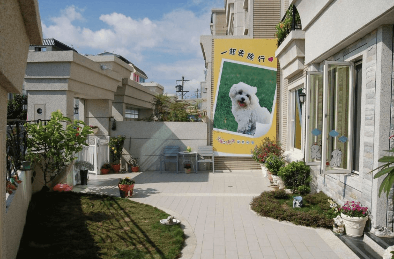 狗快樂寵物民宿戶外的陽光庭院入口。石板鋪設步道兩側種植有充滿生機的綠色植栽與盆栽。民宿米白色外牆上掛著醒目的黃色看板,上面印有可愛的比熊犬照片以及「一起去旅行」與「狗快樂寵物民宿」字樣。