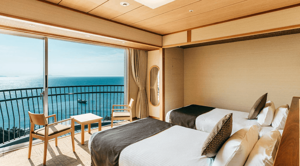 玄界灘龜之井飯店 (KAMENOI HOTEL GENKAINADA)2.png 飯店海景房實景。室內採用淺色木質調設計，配有兩張單人床。落地窗前設有木質桌椅休息區，窗外即是廣闊湛藍的海洋與晴朗天空。陽光灑進室內，展現出明亮開闊的度假感。