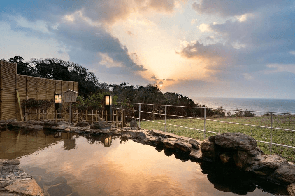 玄界灘龜之井飯店 (KAMENOI HOTEL GENKAINADA)3.jpg 飯店的露天風呂（溫泉）。由石頭砌成的溫泉池正對著大海，池水倒映著夕陽餘暉與雲彩。岸邊設有日式木欄杆與石燈籠，背景是落日沒入海平面的壯麗自然景觀。