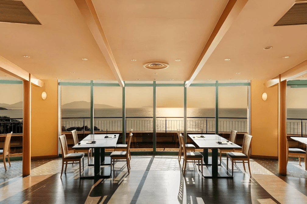 玄界灘龜之井飯店 (KAMENOI HOTEL GENKAINADA)4.png 飯店餐廳內景。餐廳擁有大面積的落地景觀窗，正對著玄界灘的夕陽美景，金色的陽光灑滿整個用餐空間。黑色長桌與木質靠背椅排列整齊，環境優雅大方，讓房客能一邊用餐一邊欣賞日落海景。