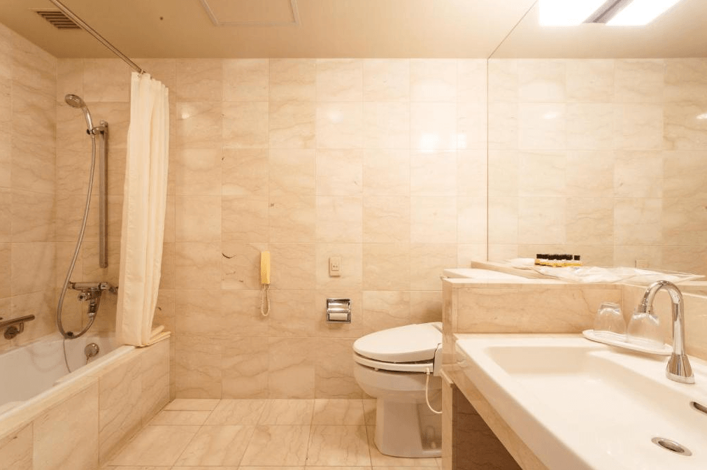 門司港普樂美雅酒店 (Premier Hotel Mojiko)3.jpg 飯店客房衛浴實景。牆面與地面皆鋪設米色大理石紋磁磚，視覺感溫潤。配置有浴缸、免治馬桶與洗手台，大型鏡面延伸了空間感，整理環境乾淨且光線充足。