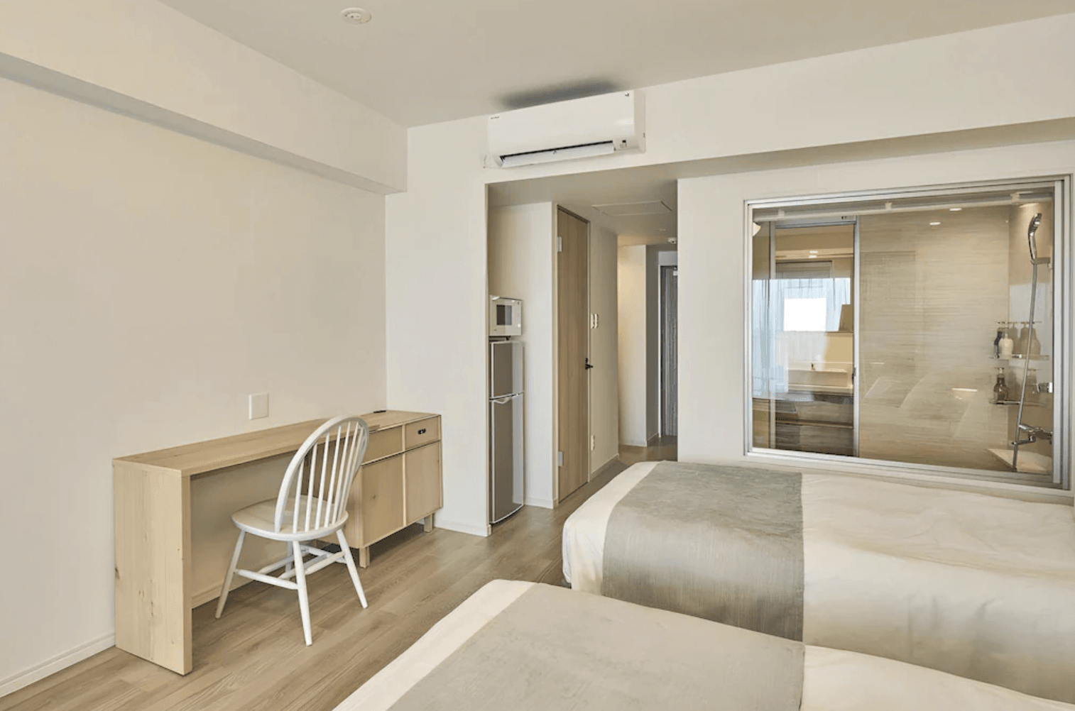 沖繩宮古島水印飯店 (Watermark Hotel Okinawa Miyakojima) 的客房內部空間。整體以純白與淺木色系為主調，左側設有長型木質書桌與白色靠背椅，後方配置了附有微波爐與雙層冰箱的簡易茶水間，右側則是通透的透明玻璃隔間衛浴。